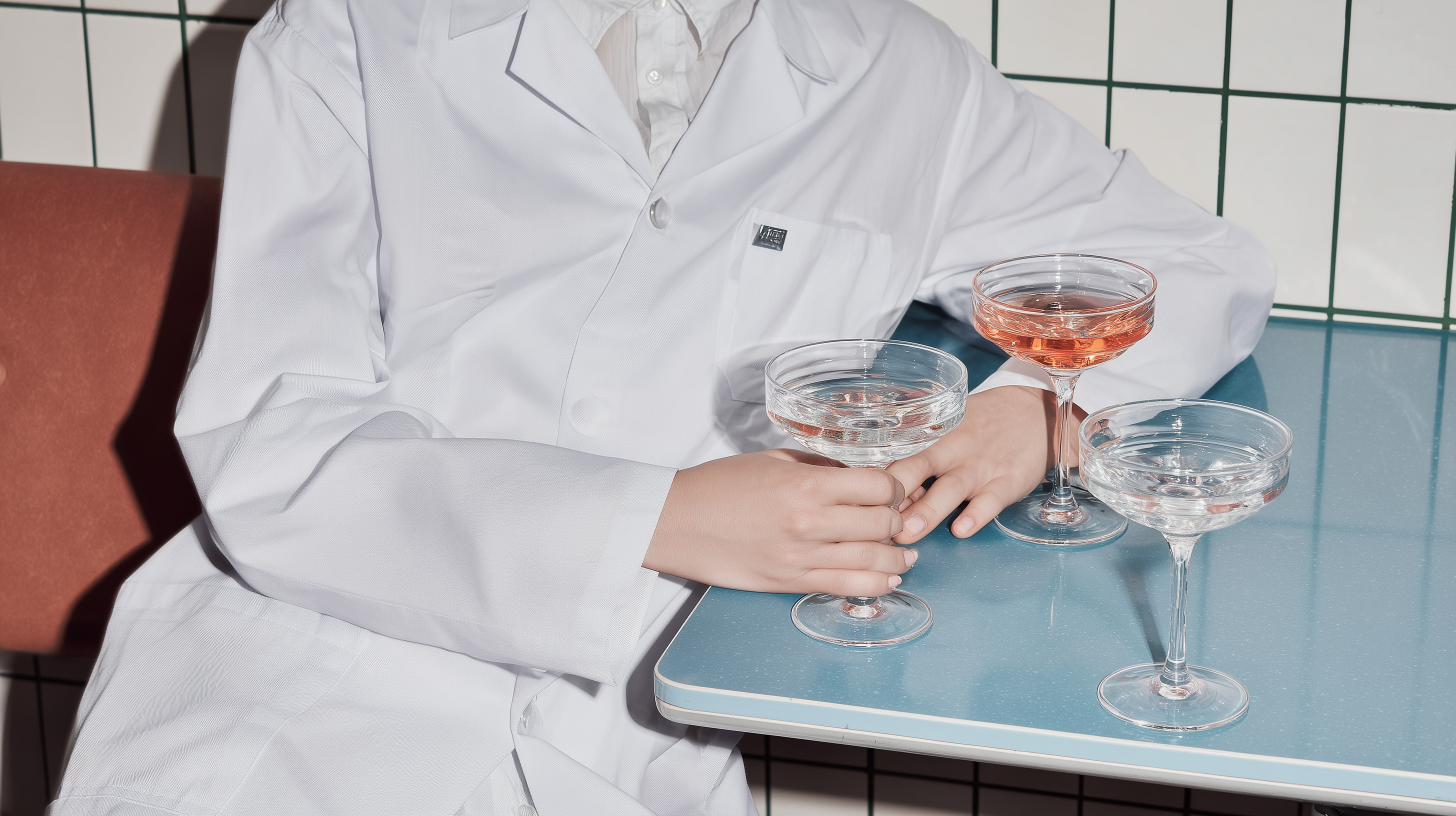 Scientist in a white coat holding three cocktail glasses with drinks on a table in front of tiled wall.